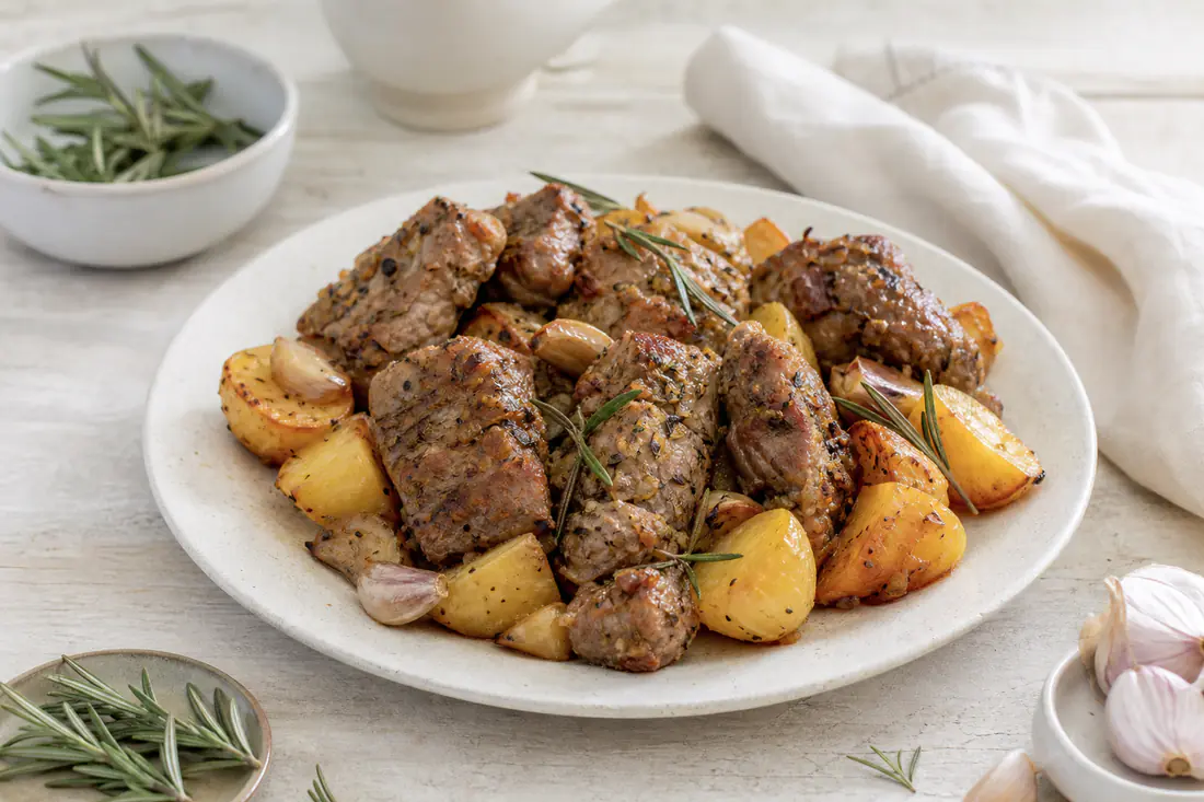 Pezzi di agnello arrosto dorati con rametti di rosmarino e spicchi d’aglio, serviti su un piatto con patate