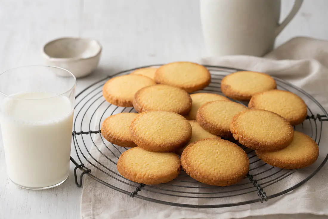 Biscotti rotondi di frolla dorati con il bordo croccante, appoggiati su una griglia di raffreddamento accanto a una tazza di latte