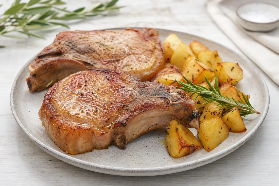 Due braciole di maiale con l’osso dorate e lucide, con il bordo di grasso croccante, servite con patate al forno e rosmarino