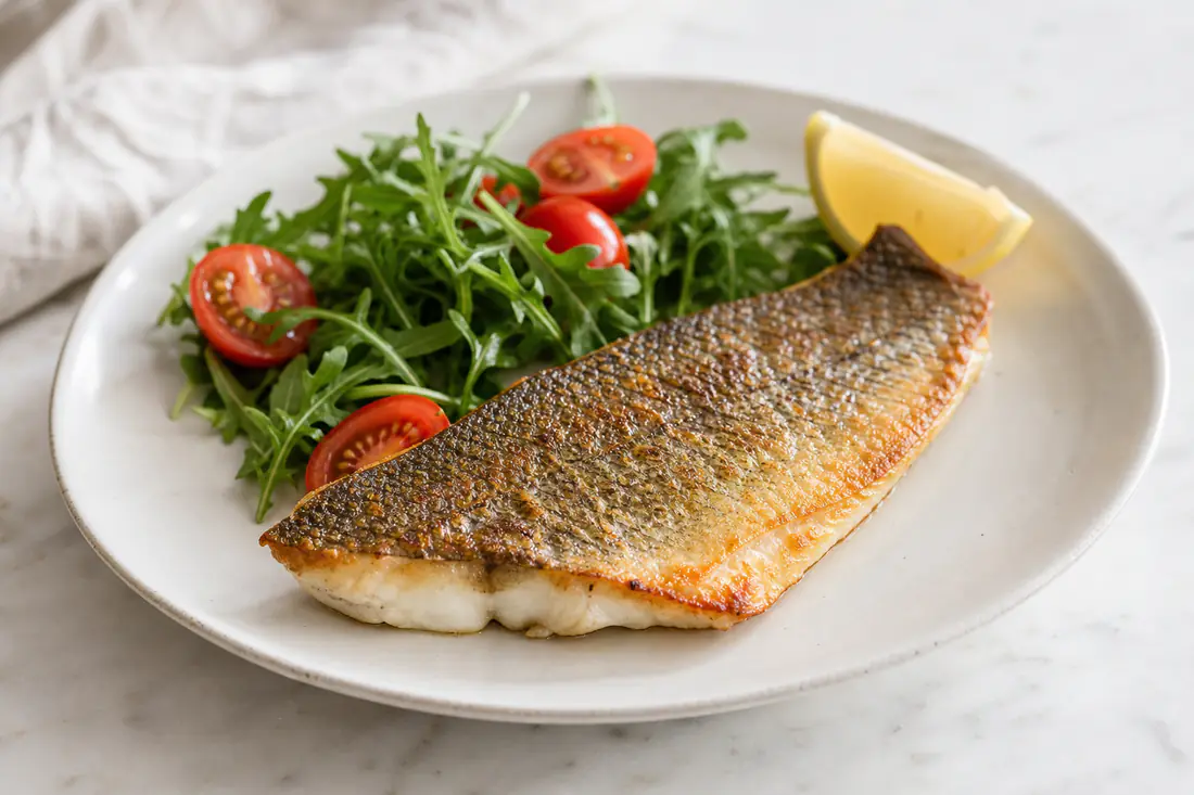 Filetto di branzino con la pelle dorata e croccante servito su un piatto con rucola, pomodorini e uno spicchio di limone
