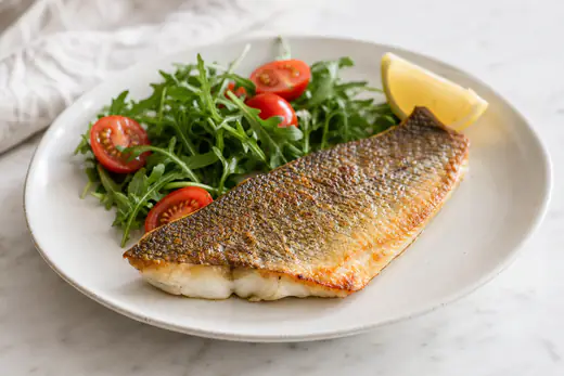 Filetto di branzino con la pelle dorata e croccante servito su un piatto con rucola, pomodorini e uno spicchio di limone