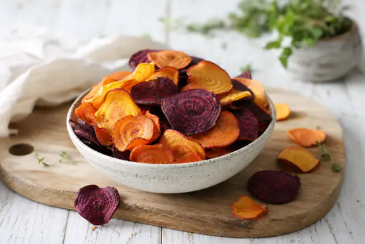 Chips croccanti di carota arancione, patata dolce e barbabietola viola in una ciotola, con fette sottili sparse su un tagliere