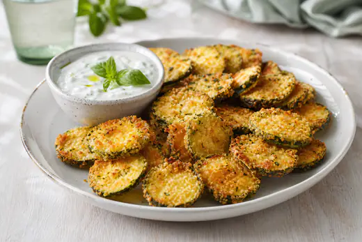 Fette rotonde di zucchina impanate e dorate con pangrattato e parmigiano, accanto a una ciotolina di salsa allo yogurt