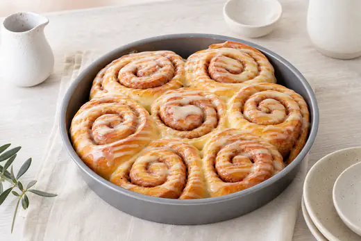 Sei cinnamon rolls a girella dorati in una tortiera tonda, con glassa bianca che cola sopra e cannella visibile tra le spirali