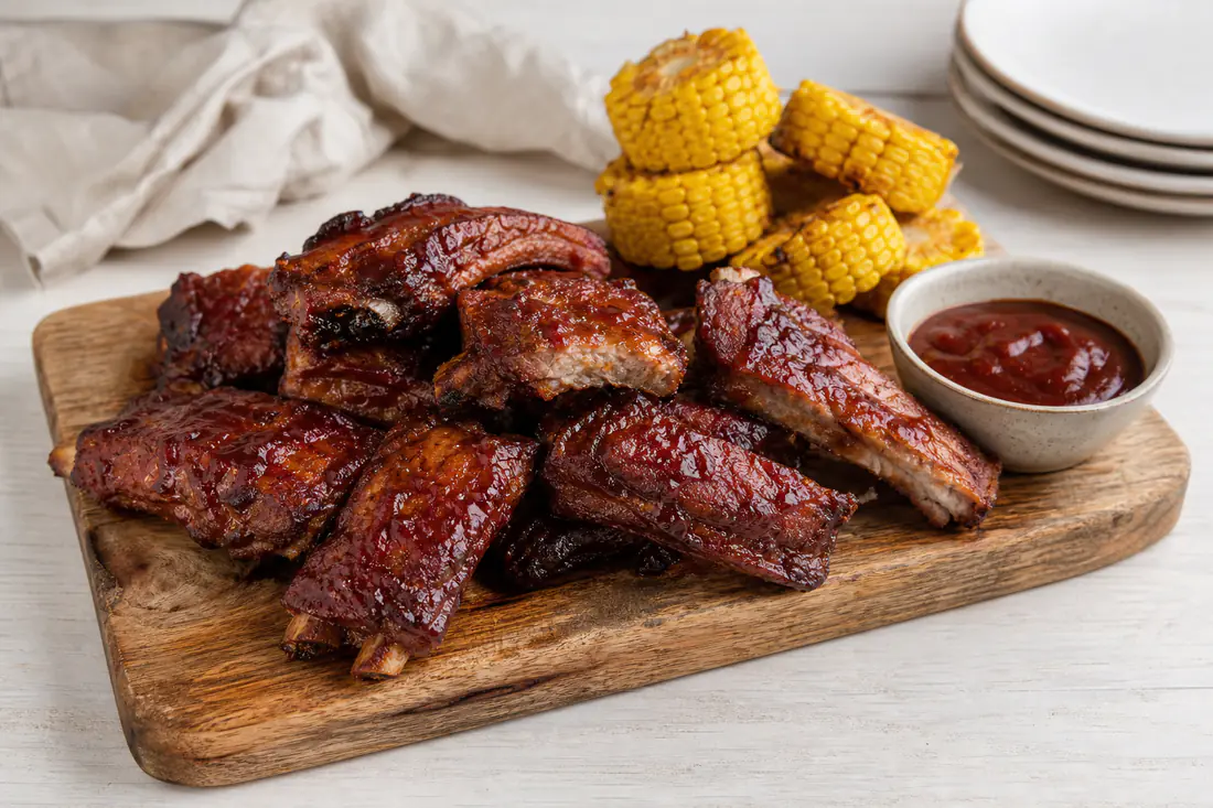 Costine di maiale glassate con salsa barbecue su un tagliere di legno, tagliate a porzioni con le ossa in vista, con mais e salsa a lato