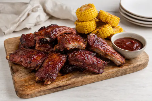 Costine di maiale glassate con salsa barbecue su un tagliere di legno, tagliate a porzioni con le ossa in vista, con mais e salsa a lato