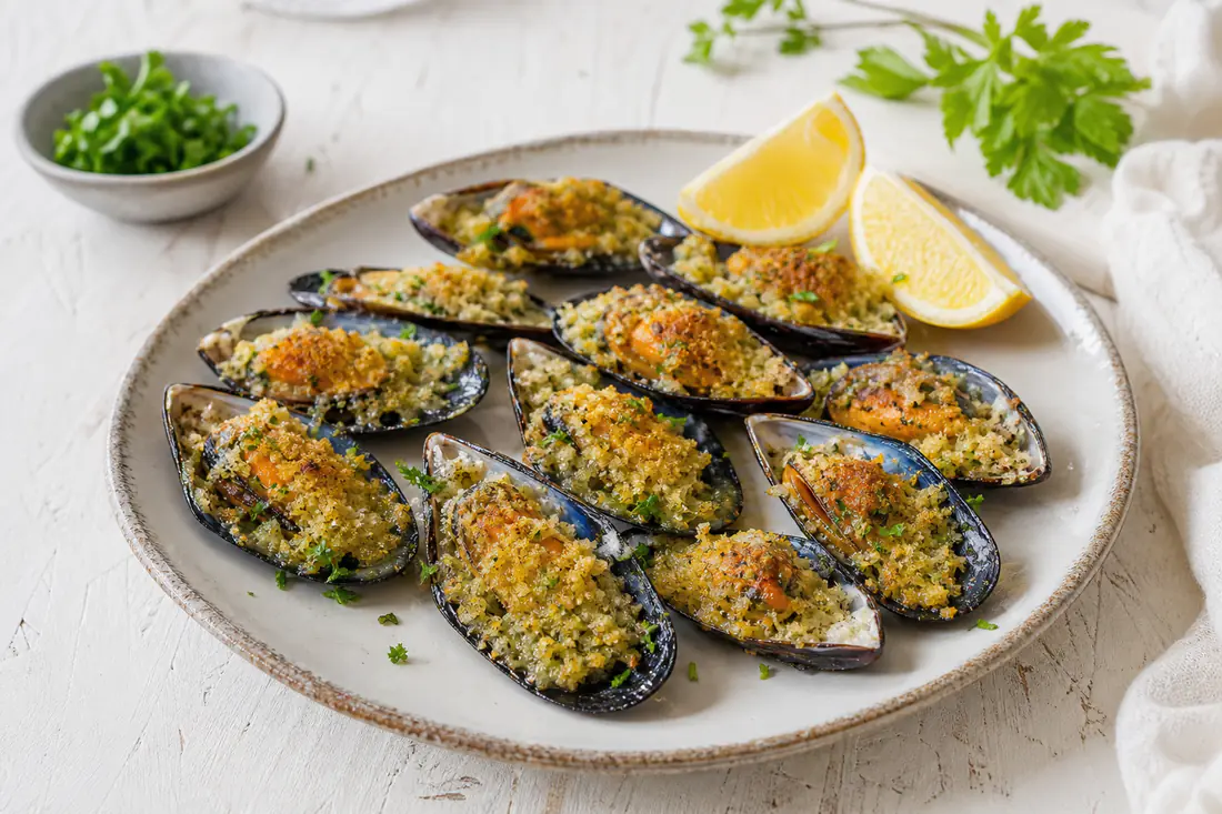 Cozze gratinate su mezza conchiglia nera, con una crosticina dorata di pangrattato e prezzemolo, disposte su un piatto con spicchi di limone