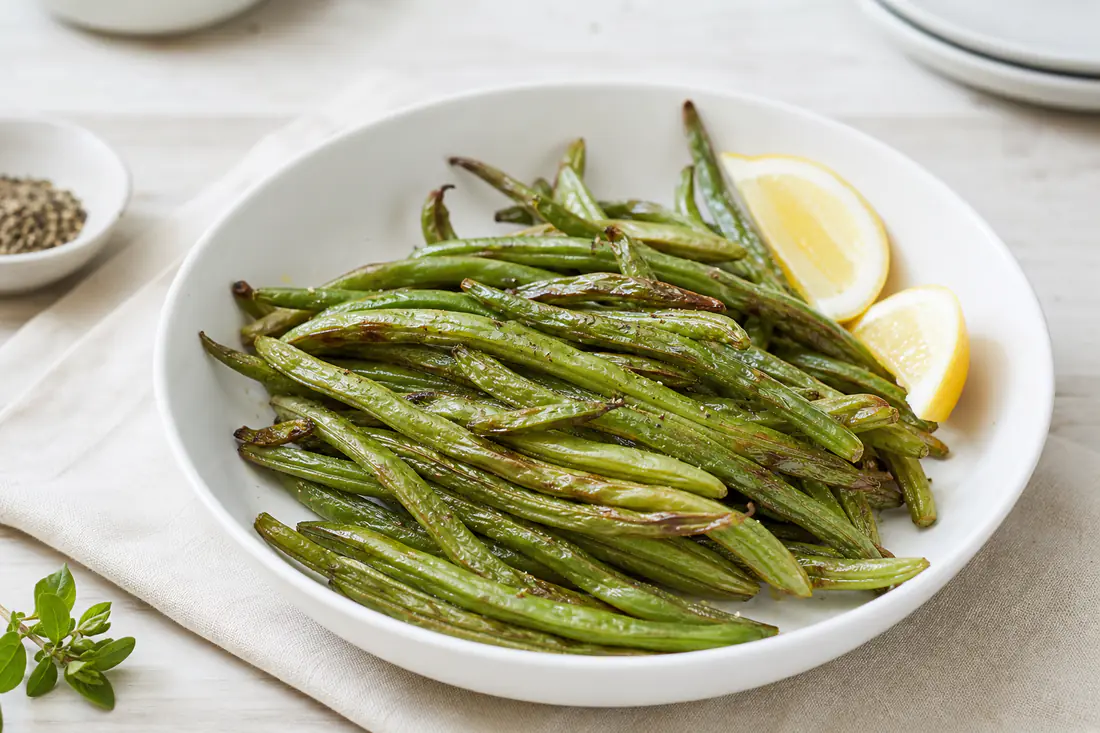 Fagiolini verdi arrostiti con le punte dorate e la buccia leggermente grinzosa, serviti in una ciotola con spicchi di limone