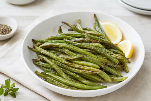 Fagiolini verdi arrostiti con le punte dorate e la buccia leggermente grinzosa, serviti in una ciotola con spicchi di limone