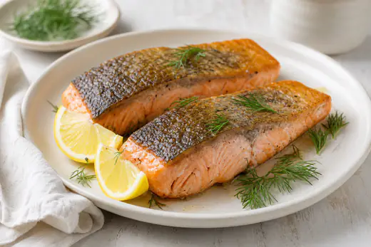 Due filetti di salmone con la pelle croccante e la polpa rosata, serviti su un piatto con spicchi di limone e rametti di aneto