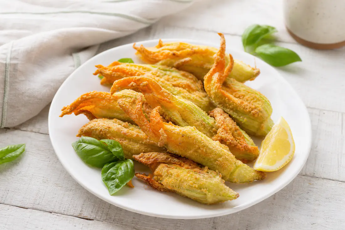 Fiori di zucca ripieni di ricotta, impanati e dorati, in un piatto bianco con un ciuffo di basilico e uno spicchio di limone