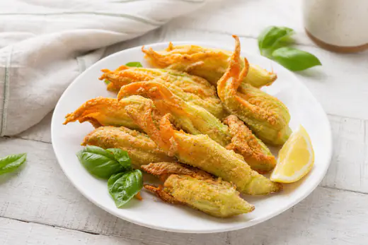 Fiori di zucca ripieni di ricotta, impanati e dorati, in un piatto bianco con un ciuffo di basilico e uno spicchio di limone