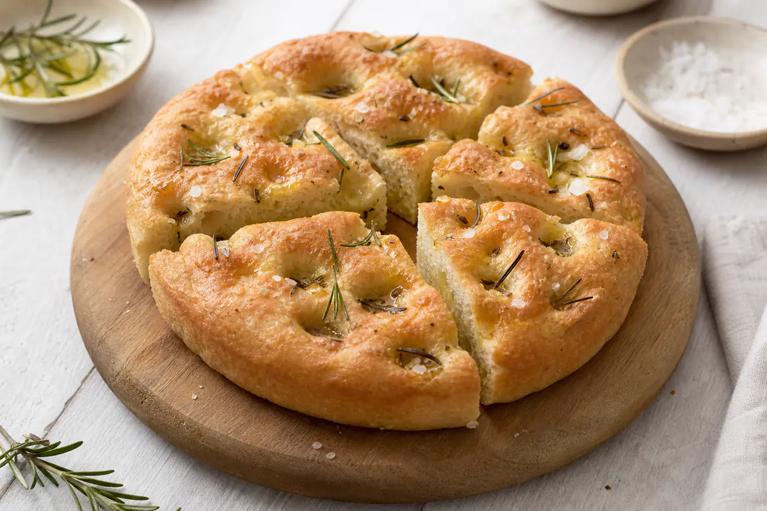 Focaccia rotonda alta e dorata con i classici buchi, olio d’oliva, sale grosso e rametti di rosmarino, tagliata a spicchi su un tagliere
