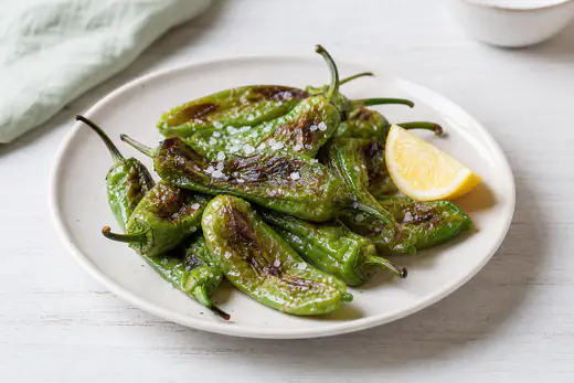 Friggitelli verdi interi con la pelle increspata e leggermente scurita, cosparsi di sale grosso, in un piatto bianco con uno spicchio di limone
