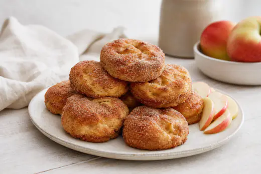 Frittelle di mele tonde e dorate rotolate nello zucchero e cannella, impilate su un piatto con mele a fette accanto