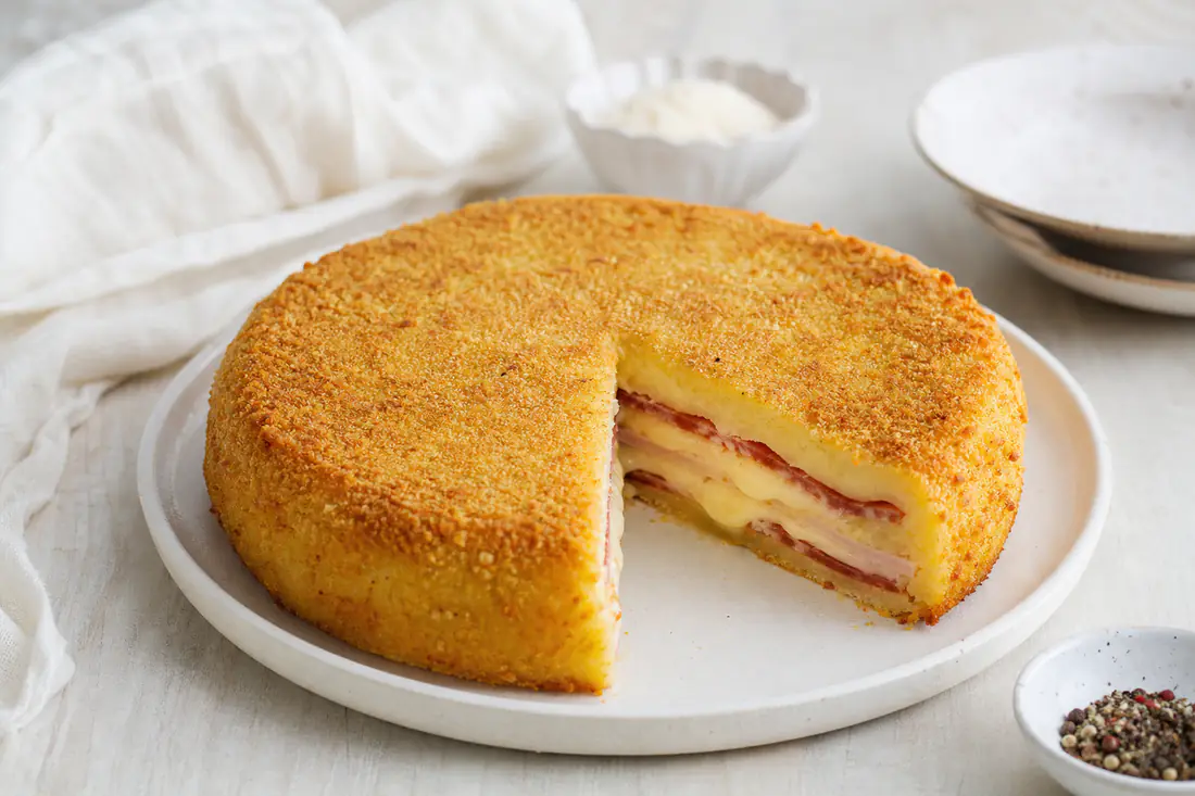 Gateau di patate rotondo sformato su un piatto, con crosta dorata di pangrattato e una fetta tagliata che mostra salame, prosciutto e provola filante