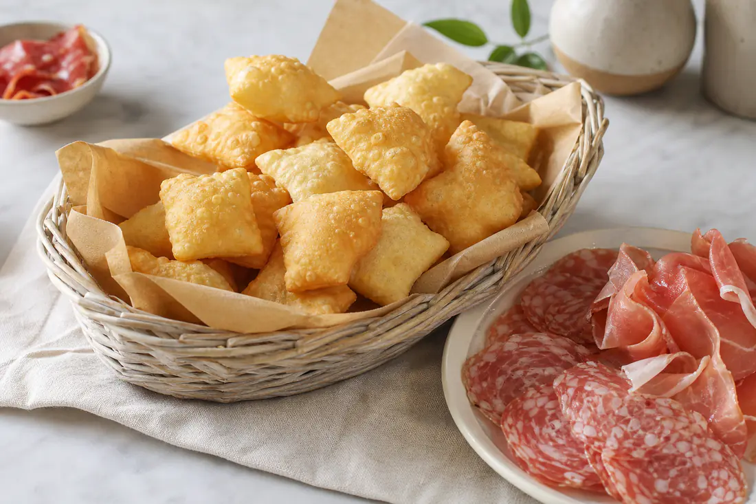 Rombi di gnocco fritto gonfi e dorati, serviti caldi in un cesto con fette di prosciutto crudo e salame