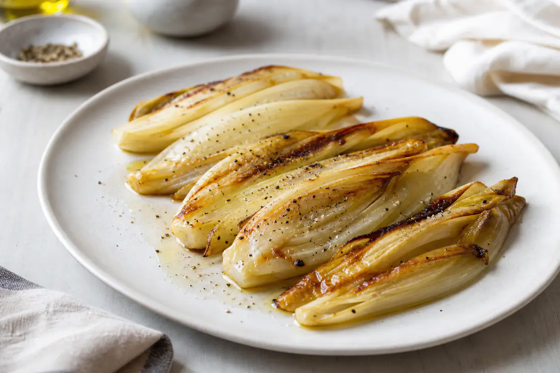 Cespi di indivia belga tagliati a metà, dorati e caramellati ai bordi, con un filo d’olio e pepe su un piatto bianco