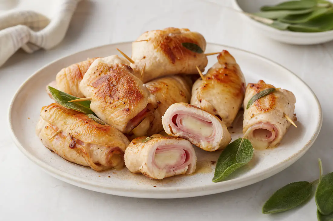 Involtini di pollo dorati ripieni di prosciutto e formaggio fuso, fermati con stuzzicadenti, uno tagliato a metà, con foglie di salvia
