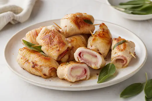 Involtini di pollo dorati ripieni di prosciutto e formaggio fuso, fermati con stuzzicadenti, uno tagliato a metà, con foglie di salvia