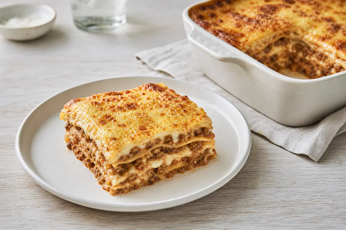 Porzione di lasagna alla bolognese con strati visibili di ragù e besciamella e crosticina dorata di parmigiano, in una piccola teglia quadrata