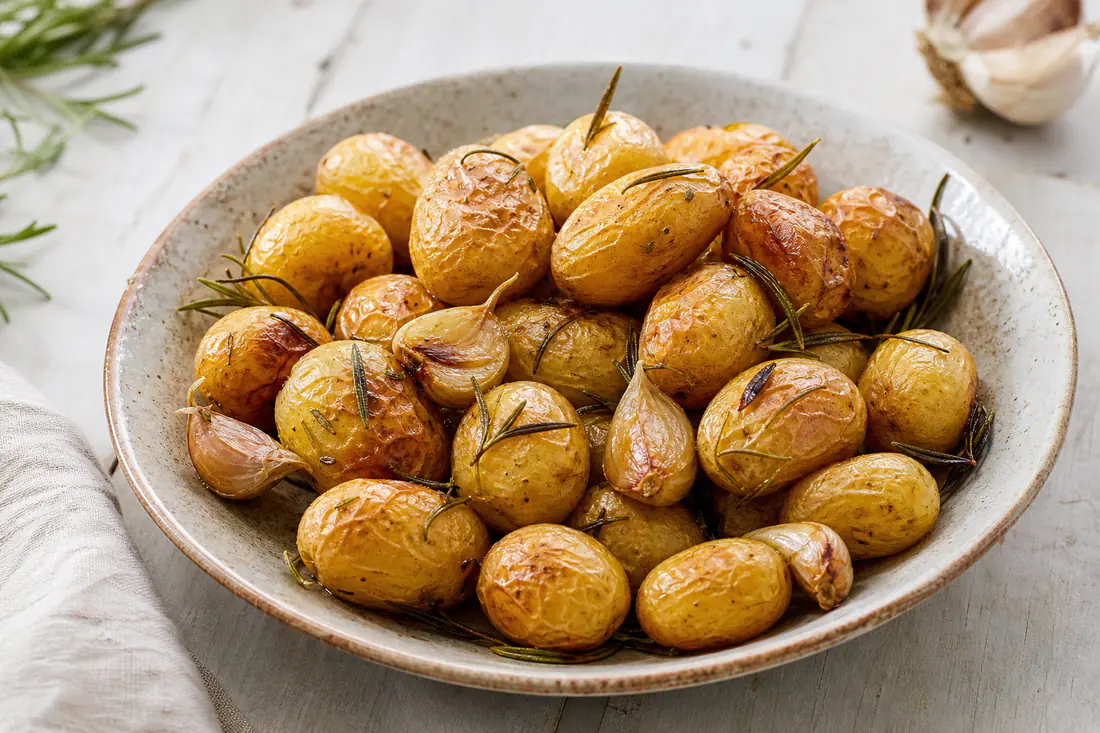 Piccole patate novelle intere con la buccia dorata e rugosa, con rametti di rosmarino e spicchi d’aglio in camicia