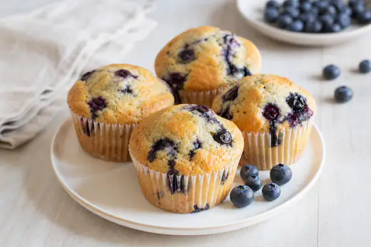Muffin ai mirtilli dorati con mirtilli viola visibili sulla cupola, serviti su un piatto bianco con una manciata di mirtilli freschi