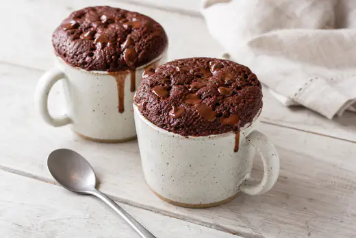 Due tazze di ceramica con mug cake al cioccolato lievitato sopra il bordo, gocce di cioccolato fuse e un cucchiaino accanto
