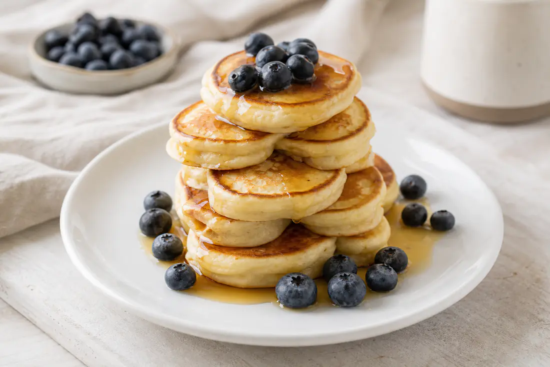 Pila di mini pancake alti e dorati con sciroppo d’acero e frutti di bosco, serviti su un piatto bianco