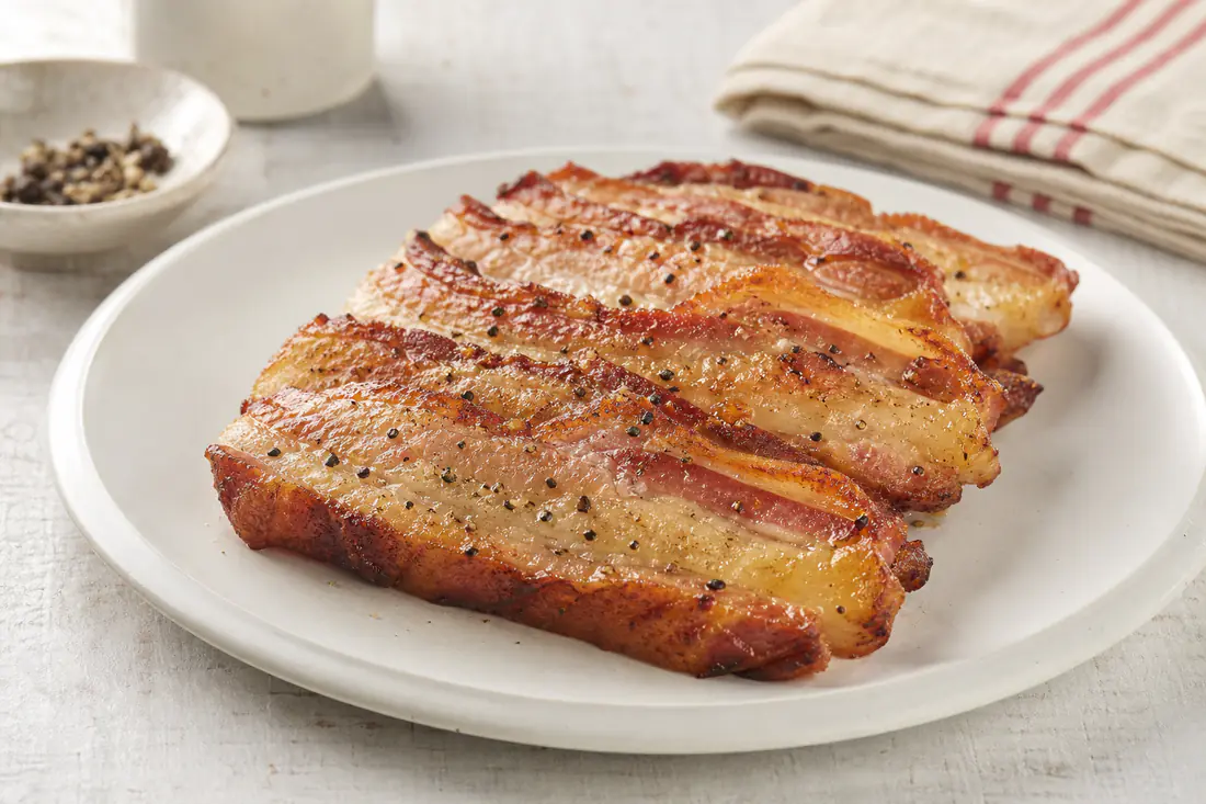 Fette spesse di pancetta arrostita, dorate e croccanti con venature di grasso, su un piatto bianco con una macinata di pepe