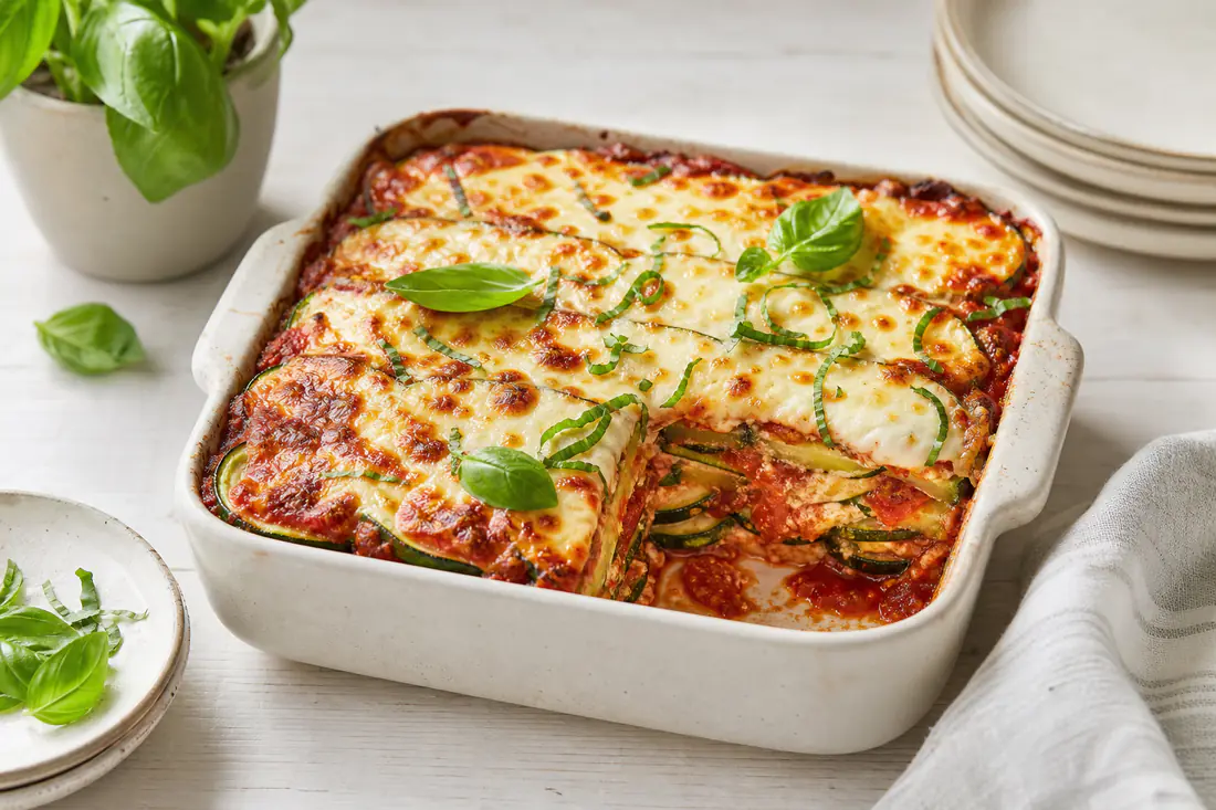 Parmigiana di zucchine in una teglia quadrata, con strati di zucchine verdi, sugo di pomodoro e mozzarella dorata, foglie di basilico