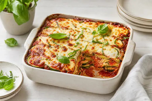 Parmigiana di zucchine in una teglia quadrata, con strati di zucchine verdi, sugo di pomodoro e mozzarella dorata, foglie di basilico