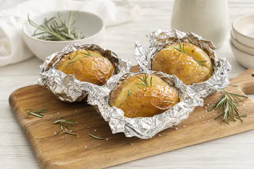 Patate intere cotte al cartoccio con la stagnola aperta, buccia dorata, rametti di rosmarino e sale grosso, su un tagliere di legno