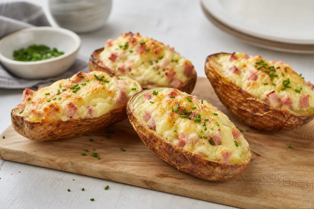 Mezze patate al forno con buccia dorata, ripiene di crema di patate, prosciutto cotto e scamorza gratinata, su un tagliere con erba cipollina