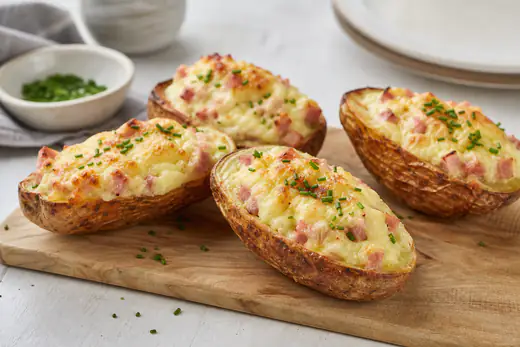 Mezze patate al forno con buccia dorata, ripiene di crema di patate, prosciutto cotto e scamorza gratinata, su un tagliere con erba cipollina