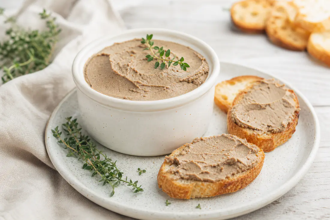 Paté di fegato liscio e cremoso in una ciotolina, spalmato su crostini di pane tostato con rametti di timo