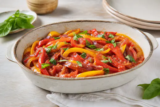 Peperonata di peperoni rossi e gialli con cipolla e pomodori in una teglia di ceramica, con foglie di basilico fresco
