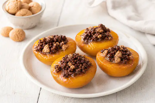 Quattro metà di pesca gialla ripiene di amaretti sbriciolati e cacao, dorate e lucide, servite su un piatto