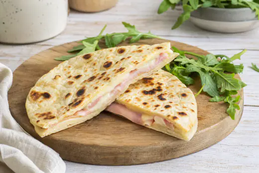 Piadina farcita piegata a mezzaluna, dorata con qualche bolla scura, tagliata a metà per mostrare prosciutto cotto e formaggio filante, con rucola accanto
