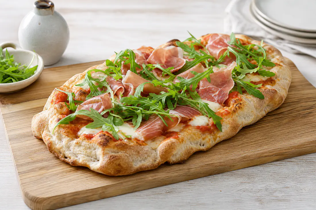 Pinsa ovale alta e alveolata con mozzarella fusa, pomodoro, prosciutto crudo e rucola fresca, su un tagliere di legno
