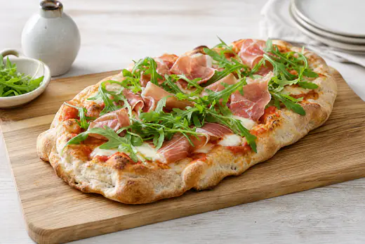 Pinsa ovale alta e alveolata con mozzarella fusa, pomodoro, prosciutto crudo e rucola fresca, su un tagliere di legno