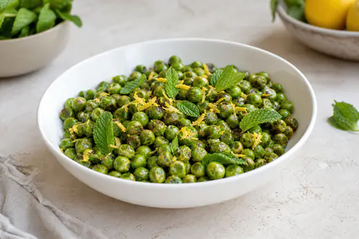 Piselli verdi brillanti, leggermente arrostiti e lucidi di olio, con scorza di limone e foglie di menta in una ciotola bianca