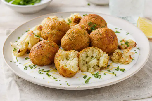 Polpette di cavolfiore dorate con pezzetti chiari di cavolfiore visibili nella polpetta spezzata, servite con salsa allo yogurt e prezzemolo