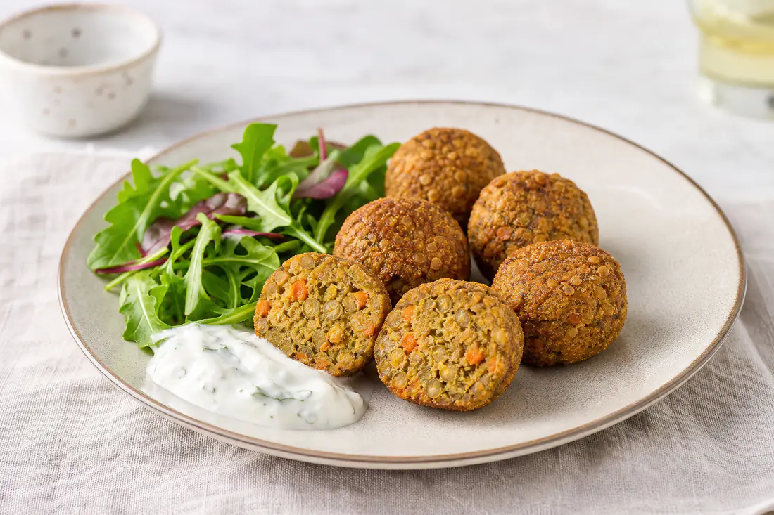 Polpette di lenticchie scure e dorate con una polpetta tagliata a metà, su un piatto con insalata verde e salsa di yogurt
