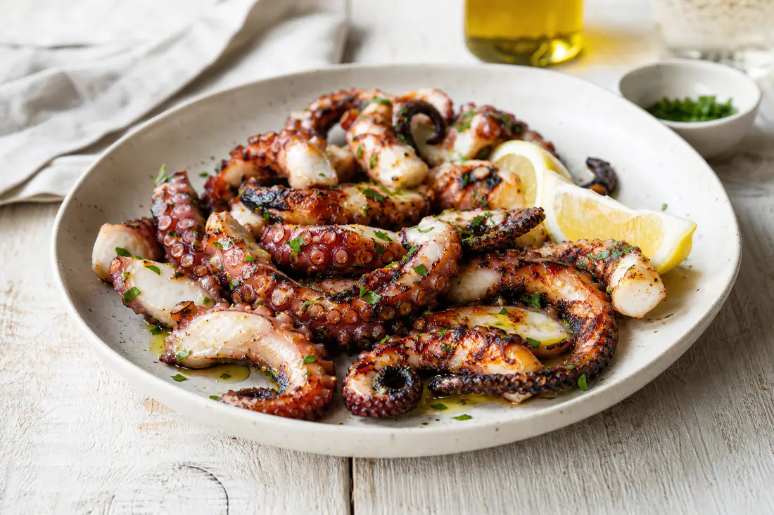 Tentacoli di polpo arrostiti con punte croccanti e pelle scura, serviti su un piatto con olio, prezzemolo e spicchi di limone