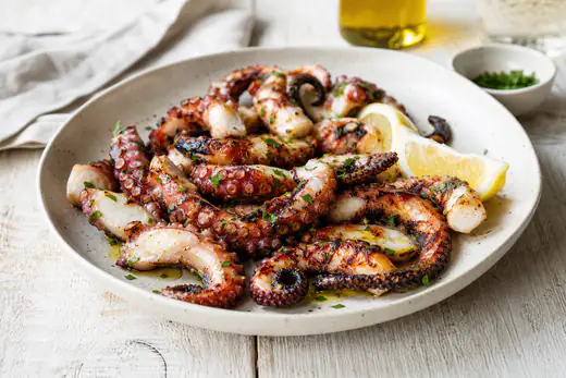 Tentacoli di polpo arrostiti con punte croccanti e pelle scura, serviti su un piatto con olio, prezzemolo e spicchi di limone