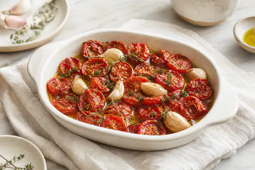 Pomodorini rossi tagliati a metà, raggrinziti e lucidi di olio, con spicchi di aglio e rametti di timo in una piccola teglia