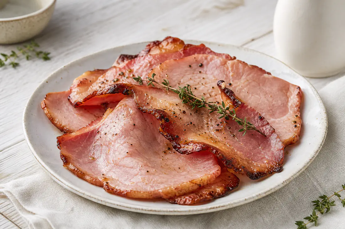 Fette di prosciutto cotto dorate e croccanti ai bordi, disposte su un piatto con pepe nero e rametti di timo