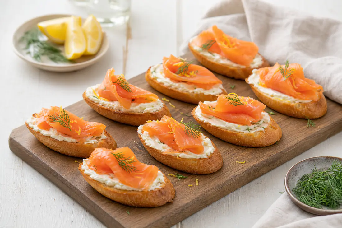 Crostini dorati con formaggio fresco e fette di salmone affumicato, decorati con aneto e scorza di limone, serviti su un tagliere di legno