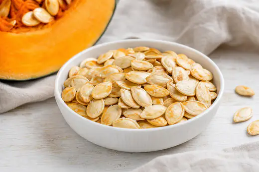 Semi di zucca con il guscio chiaro, tostati e leggermente dorati, in una ciotola bianca accanto a una fetta di zucca arancione